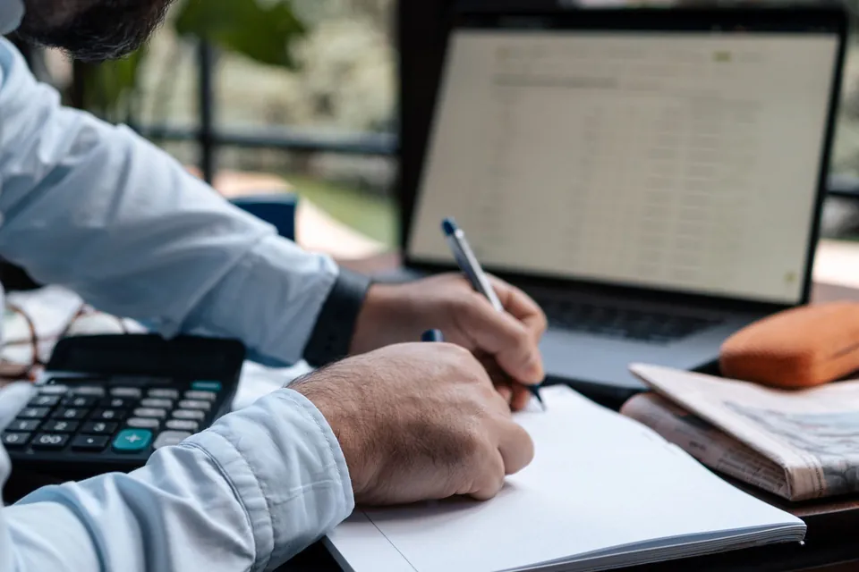 Homem anota contas ao lado do notebook e da calculadora