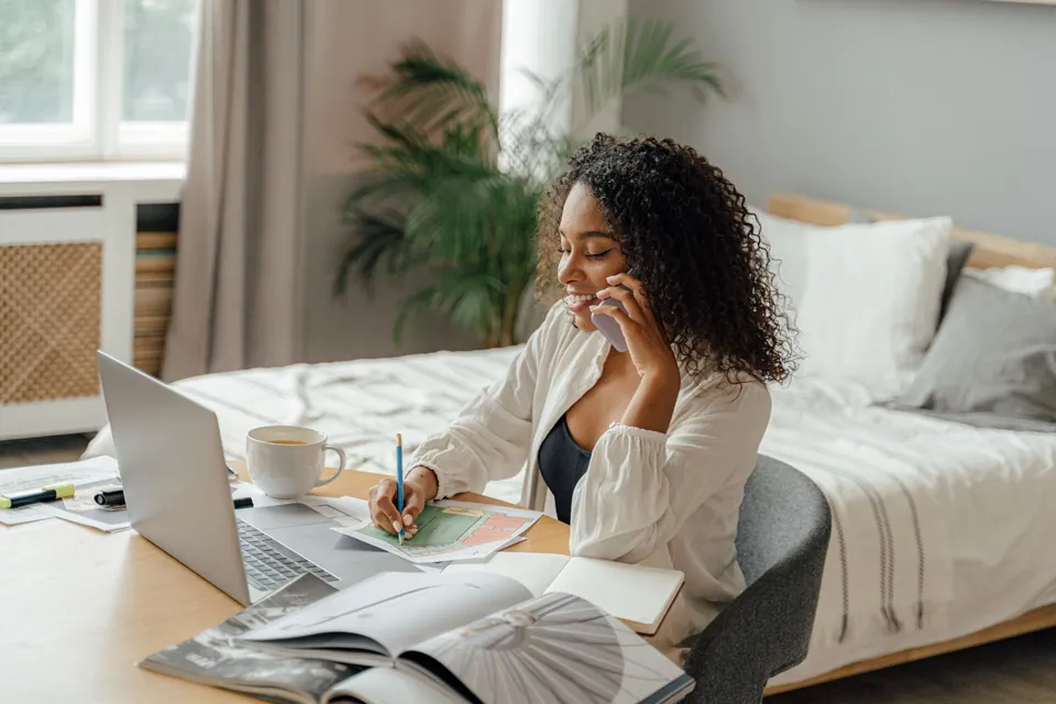 Mulher fala ao telefone e faz anotações à mesa, com notebook e papéis