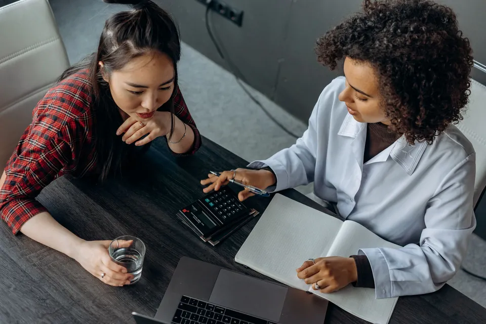 Duas pessoas conferem números no notebook e em papéis