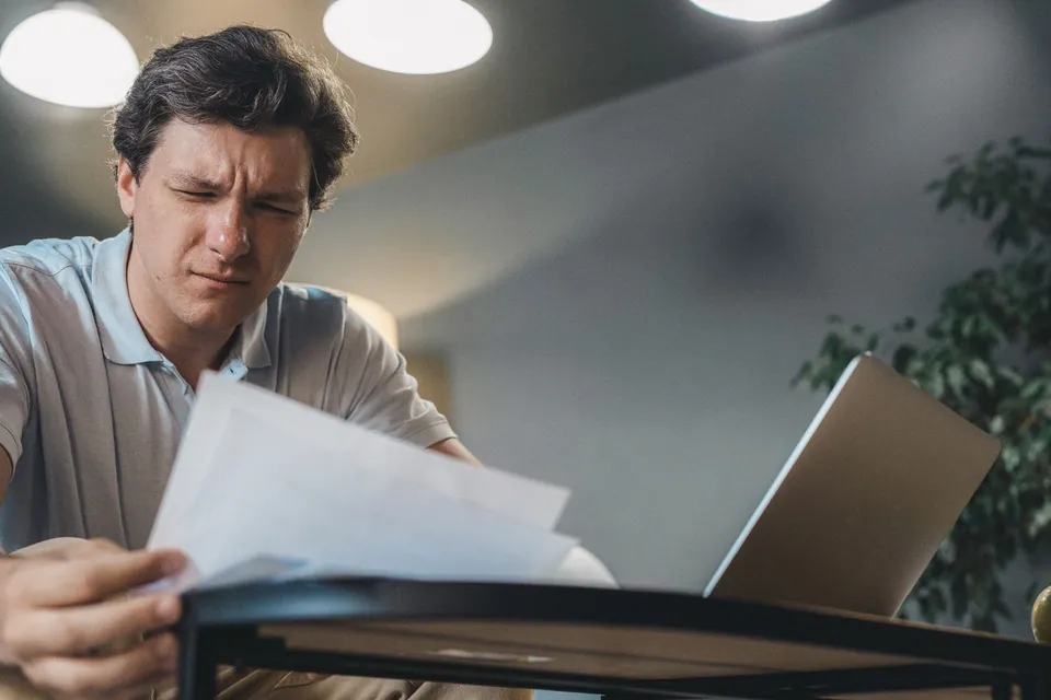 Homem confere documentos à mesa, ao lado do notebook
