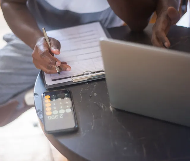 Mão anota em uma prancheta ao lado do notebook e da calculadora