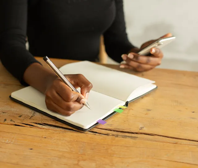 Mulher escreve em um caderno segurando o celular