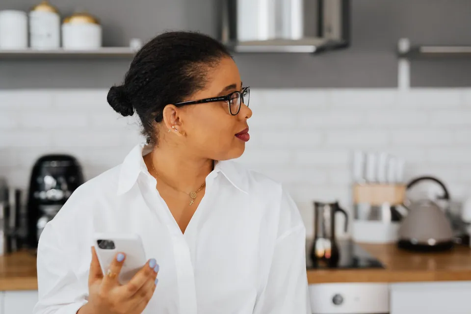 Mulher com o celular na mão olha de lado, atenta, na cozinha de casa