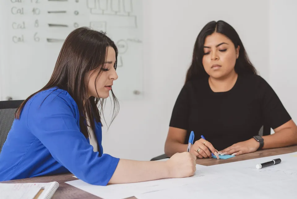 Duas mulheres conversam à mesa enquanto uma delas faz anotações