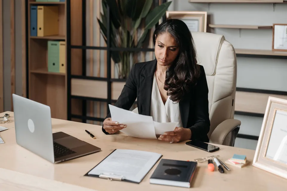 Mulher confere documentos à mesa do próprio escritório, com o notebook ao lado