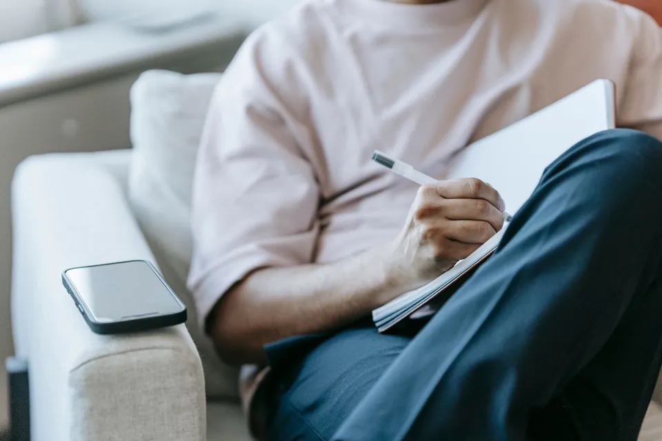 Homem anota em um caderno com o celular ao lado, sentado no sofá de casa