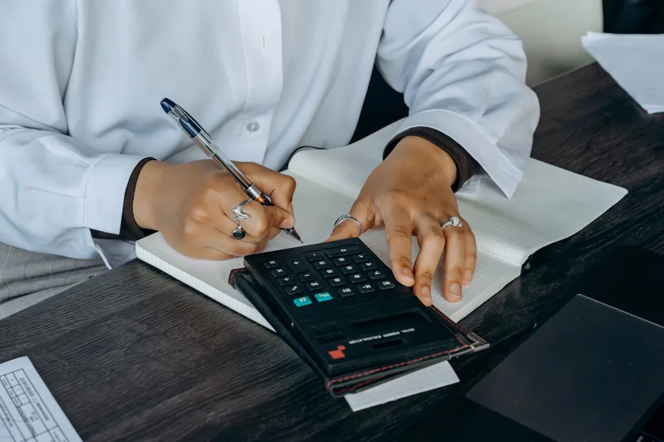 Mãos anotam números num caderno ao lado de papéis impressos e de uma calculadora