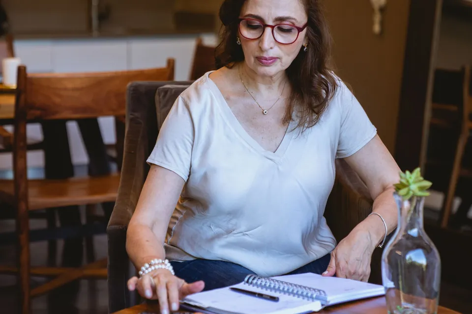 Mulher faz as contas à mesa de casa, com caderno, calculadora e celular ao lado